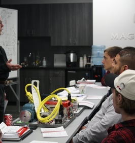 Image of older man standing, lecturing three other men, sitting down, in a room
