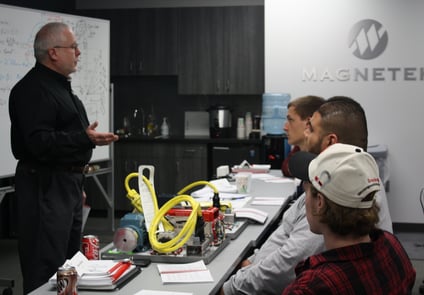 Image of older man standing, lecturing three other men, sitting down, in a room