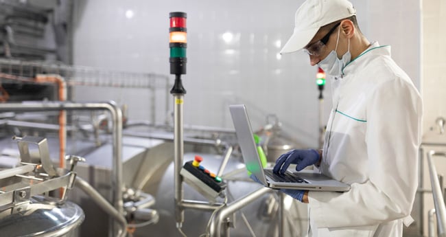 Image of Man in lab coat, goggles, hairnet, and gloves looking at computer