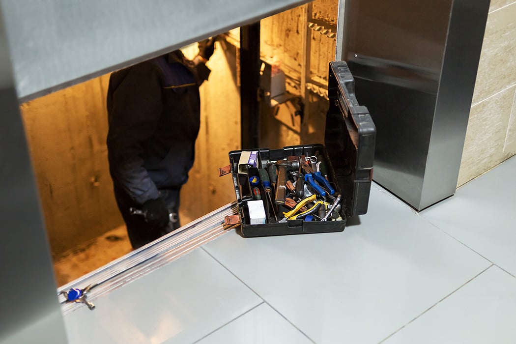 The image shows an elevator mechanic working on an elevator hoistway with an open tool case placed on the floor