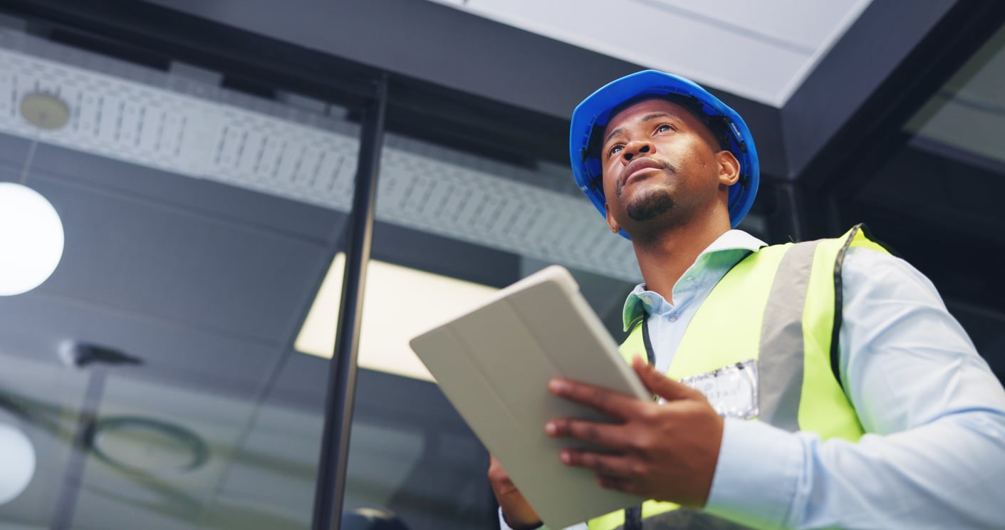 A technician wearing a blue hard hat and a high-visibility safety vest while holding a digital tablet.